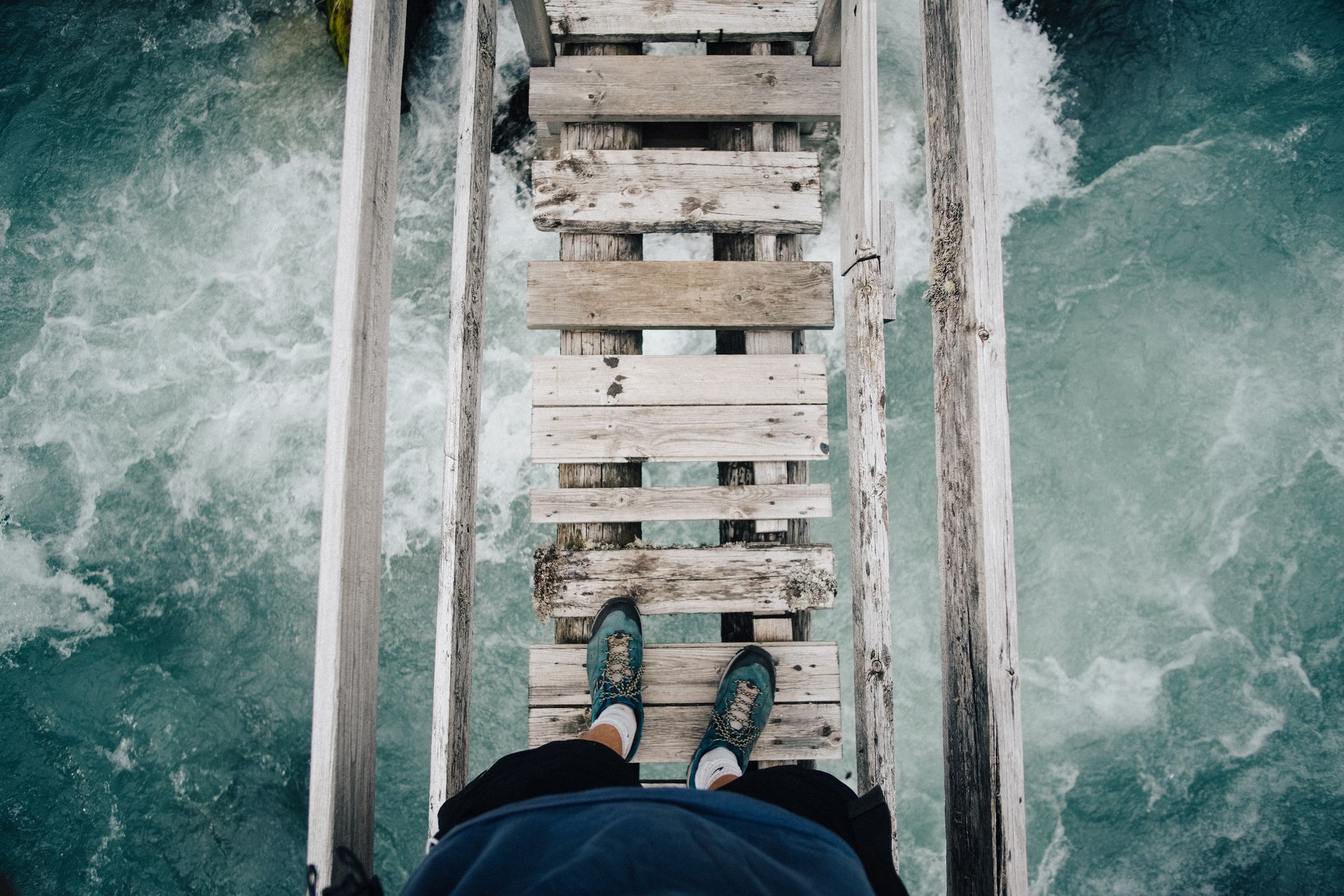 Above view of a person's feet about to cross a rickety bridge over turbulent water
