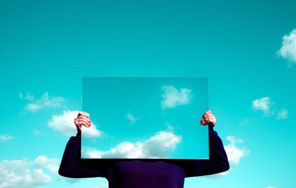 Woman holding a mirror in front of her face. The mirror is reflecting blue sky and white fluffy clouds.