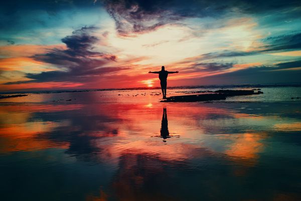 Man stood on a wet beach with arms stretched wide, silhouetted against a bright red sunset.